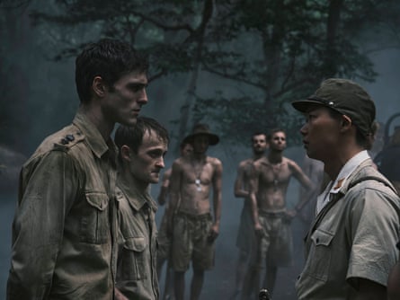 A group of young white men in ragged army uniforms standing in a darkened forest clearing, two of them facing a stern looking young Japanese man in a military uniform.