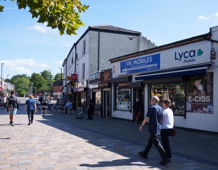 Pedestrians walk past shops including a mobile phone and e-cigarette store in Middleton town centre