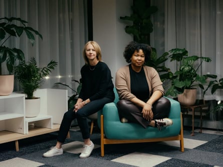 A white woman and a Black woman sit back on a teal armchair.