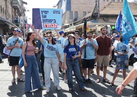 A group of young people wearing light blue T-shirts holding a sign with Hebrew writing on it, with one young woman waving her arm and yelling on a bustling street