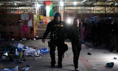 A police officer in body armor and helmet escorts a longhaired protester with hands cuffed amid debris and a Palestinian flag hanging from scaffolding.