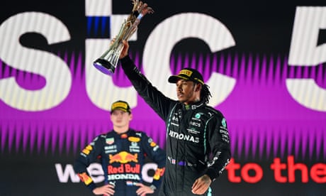 Lewis Hamilton celebrates on the podium by holding aloft the Saudi Arabian Grand Prix trophy after beating Max Verstappen, who is standing in the background with hands on hips