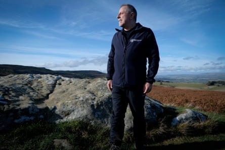 Mike Pratt stands in front of lichen-covered rocks in a moorland landscape with bracken behind him and hills in the distance. The sky above him is bright blue with small white clouds. He has short grey hair and beard and wears a dark fleece top with the wildlife trusts logo.