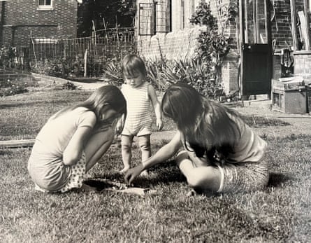A b/w photo of two girls playing a board game on a lawn with a toddler looking on