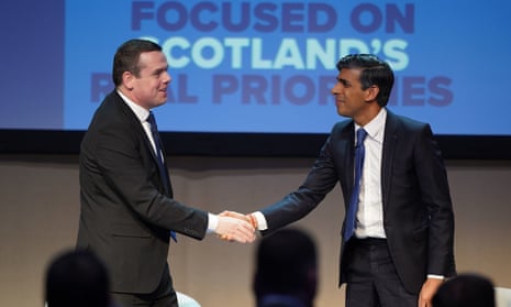 Douglas Ross and Rishi Sunak shaking hands on stage, with a message on a screen behind them saying 'Focused on Scotland's real priorities'