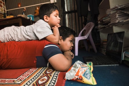 Two young Nepali boys, both overweight, lie down as they watch a screen. The smaller boy is lying on the bigger one’s back. An empty snack packet lies beside them