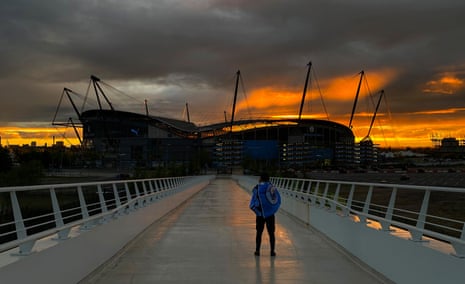 A Manchester City fan watches a golden sunset from a bridge leading to the club's home ground, the Etihad Stadium, behind which the sun is sinking.
