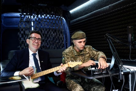 Andy Burnham playing an electric guitar while a soldier adjusts settings on a mixing desk