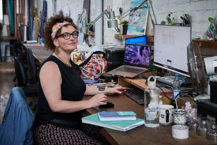 Artist Lu Campbell-Smith sits at a cluttered studio desk with computer monitors, smiling at the camera