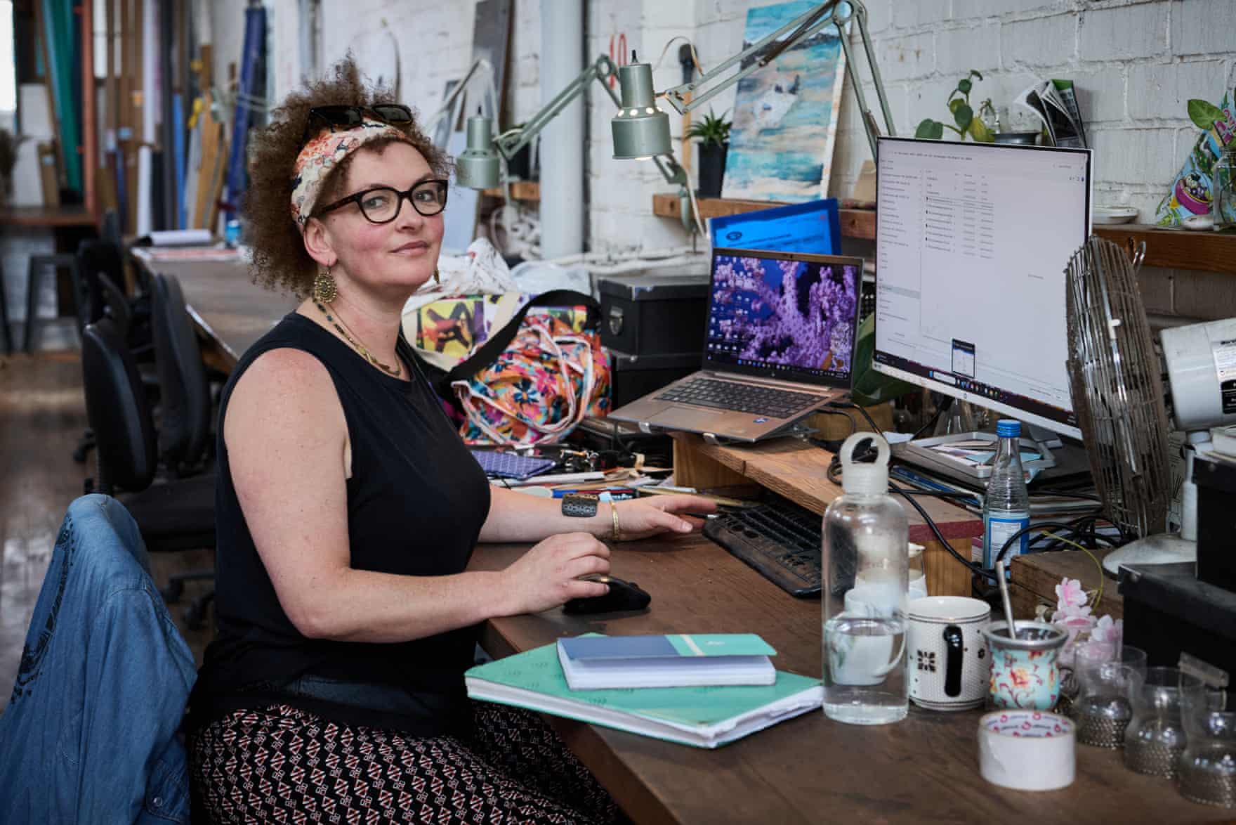 Artist Lu Campbell-Smith sits at a cluttered studio desk with computer monitors, smiling at the camera
