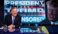 Piers Morgan laughs as he presents his TalkTV show in front of a backdrop of Donald Trump