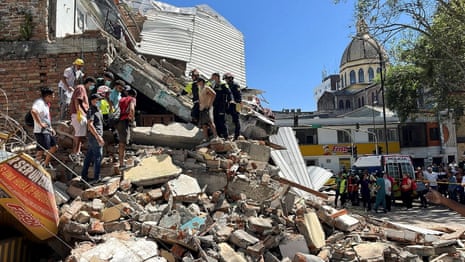 People – some in emergency uniform – dig through the rubble of a collapsed building. There is a church spire in the background.