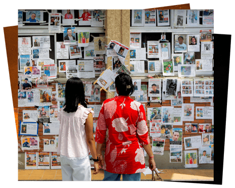Two women with their back to the camera look at a wall covered in photographs of missing people