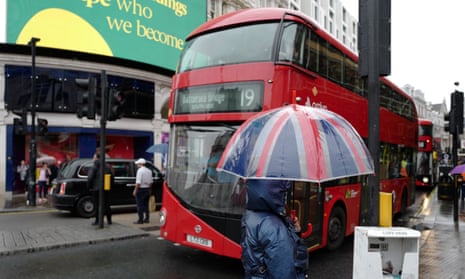A tourist at Piccadilly Circus shelters from the rain under an umbrella as a bus goes by