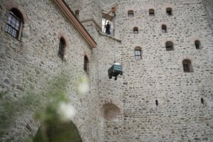 Father Isaiah stands on a balcony facing out of an old stone building, using a crane to pull up a trolly