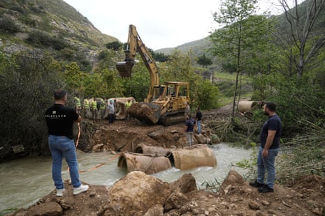 Two men setand on one side of a river that has three large muddy barrels serving as a sort of crossing. Across the way, more men and workers gather around construction machinary.
