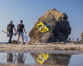 Two men carrying fishing rods and a bucket walk along a beach past a large rock with a huge pair of yellow sunglasses