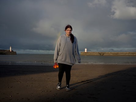 A young woman on a beach underneath a gloomy sky, holding a bag of oranges.