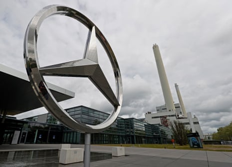 A picture of a large, freestanding Mercedes three-pointed star emblem against the sky, with buildings and chimneys of a large factory visible behind
