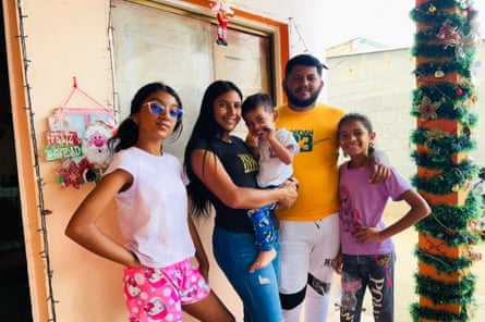 Smiling family in well-lit room decorated for Christmas, with man, woman, two teenage-ish girls, and a baby boy.
