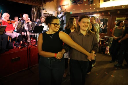 Two young women dance together with their arms crossed over. The orchestra plays on the stage behind them.