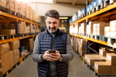 Male business owner standing in aisle of a warehouse space, boxes on shelves, holding his phone looking at screen.