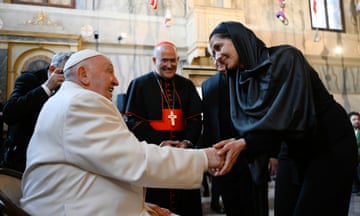 Pope Francis, seated and wearing white, holds the hands of a woman wearing a black dress and headscarf while another cleric in black and red robes looks on, smiling; they are inside an ornate stone chapel