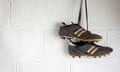A pair of muddy black football boots hang up by the laces against the white wall of a changing room.