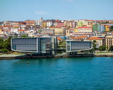 A modern building on a waterfront with water in front houses and buildings behind it.
