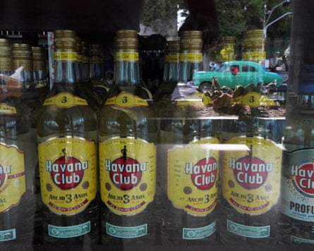 A vintage car is reflected on a window of a shop displaying bottles of Havana Club rum