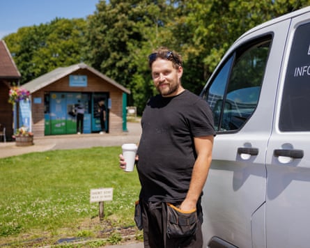 Sam stands by his white van holding a white drinks container. He wears a black T-shirt and work trousers with large pockets for tools.
