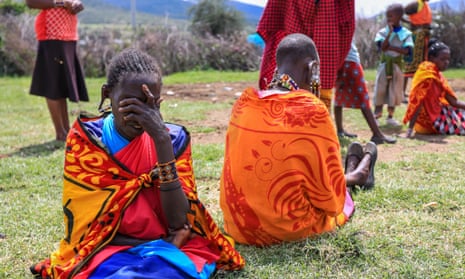 Women sit on ground, wearing bright orange traditional clothing. One woman covers her face with her hand