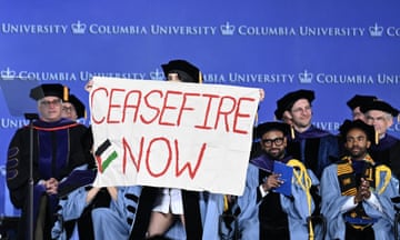 People in blue academic robes and a woman holding a sign reading 'ceasefire now' with a Palestinian flag.