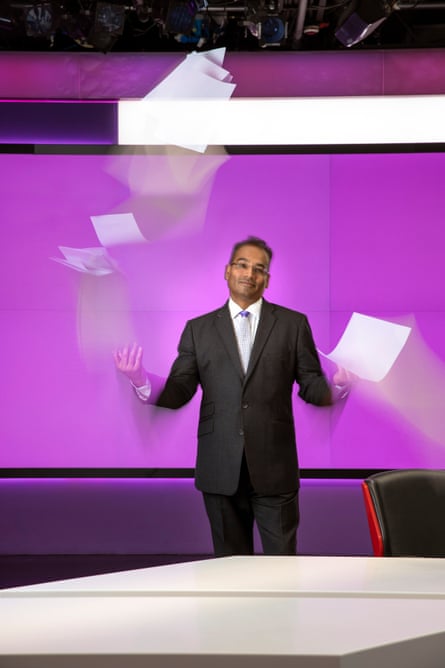 Krishnan Guru-Murthy wears black suit and white shirt; standing, shrugging and throwing paper up in the air
