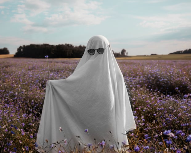 Someone dressed as a ghost with a sheet over them and sunglasses poses in a summer meadow with purple blossoms