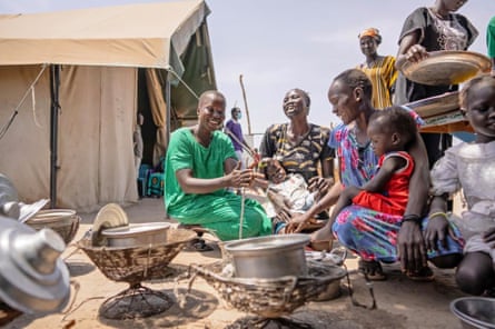 African women, some with children, sit around a cooking fire, talking and laughing, in a camp for IDPs in South Sudan.
