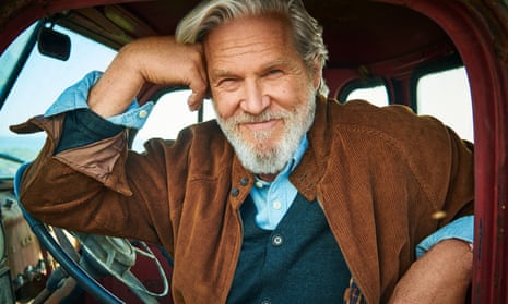 Jeff Bridges seen through the open window of a truck, leaning on the steering wheel and smiling