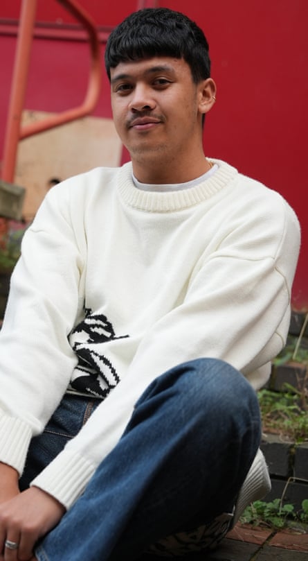 Freddie Feltham, with an earring, a slight moustache and in a crew-cut jumper and jeans, sits on brick stairs with his knee drawn up, slightly smiling
