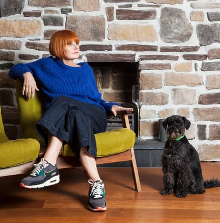 Mary Portas in an electric blue jumper, cropped wide-bottom jeans and trainers sitting cross-legged on her sofa at her home, a bare brick wall behind her and her black dog next to the sofa