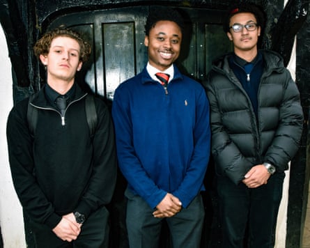 Three young men, all wearing quarter-zip sweaters, standing against the black door of a historic house