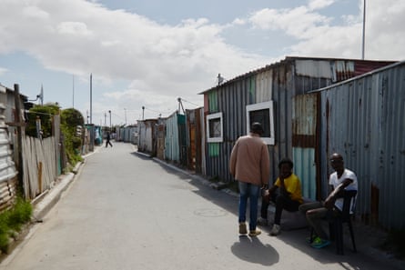A long, tarmacked road along which single-storey houses made of wood and corrugate iron are densely packed together