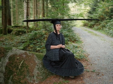 Gertraud Platschek, 2024This portrait of artist Gertraud Platschek was taken in the forest near her home in Bavaria, Germany. Seated on a rock, Platschek is seen in one of her wearable works of sculpture, a hat made from a pole covered in black fabric. The horizontality of its form contrasts with the natural environment, producing a glimmer of the surreal which British photographer Camilla Greenwell often seeks out in her work. The portrait, part documentation, part performance, was taken during a collaboration with Platschek