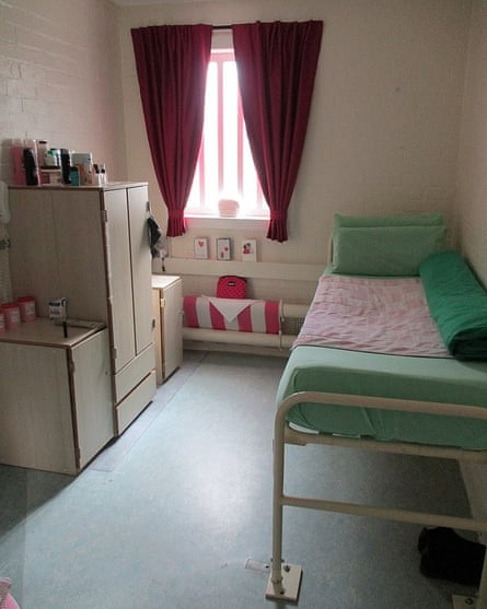 A prison cell. On the right there is a bed on a raised metal frame. In the centre, there is a barred window in the far fall with red curtains. On the left there are some storage units