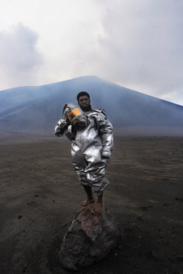 A barefoot volcanologist in a silver protective suit stands on a rock before a smoking volcano