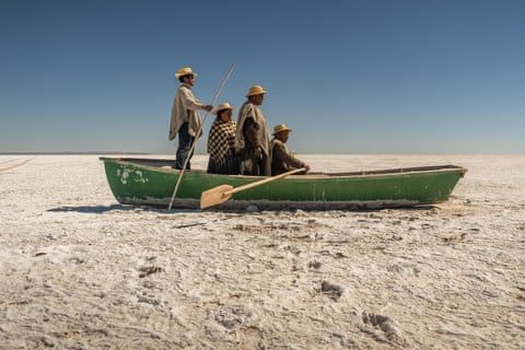 Four people in traditional Bolivian dress sit in a boat stranded on the salty desert where Lake Poopó used to be.
