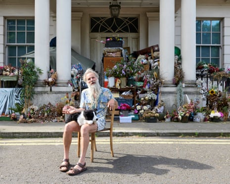 A man in a Hawaii-style shirt, cream shorts and brown sandals sits on the step of the porch of a grand pillared home, surrounded by furniture, flowers and knick-knacks