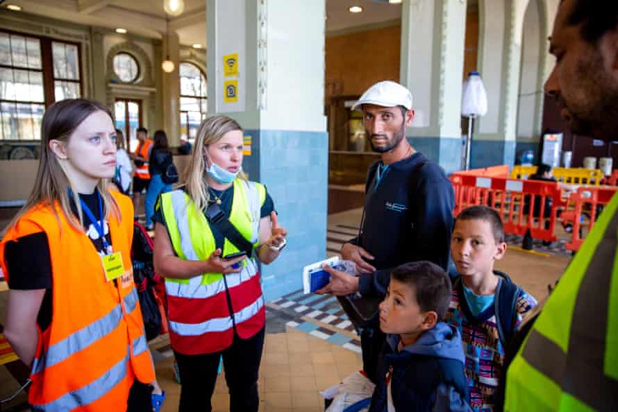 Deux jeunes femmes et un homme portant des vestes haute visibilité assistent un Rom avec deux jeunes garçons dans la gare