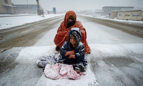 A woman and a child huddle in the middle of a snowy road. They are wrapped up in relatively thin clothing and are covered in snow