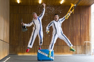 Two men in white and blue-striped overalls jump into the air while shouting and smiling as they hold a small guitar and a trombone