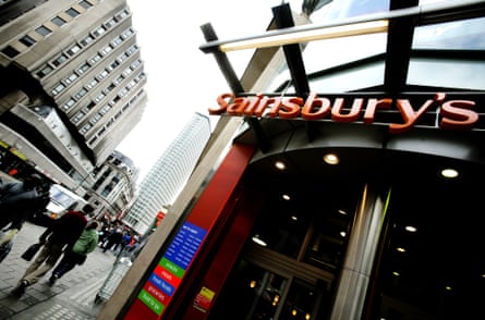 Sainsbury’s supermarket in Tottenham Court Road, London, photographed from a low angle with the doorway, sign and tall buildings in the background looming over the viewer. People are walking on the street in the background and it looks like a grey, wet and wintry day.